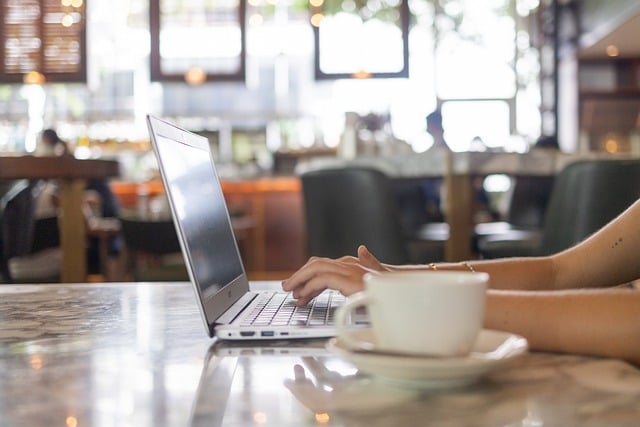 Kaffeetasse, im Hintergrund Hand einer Person am Laptop, in einem Café 